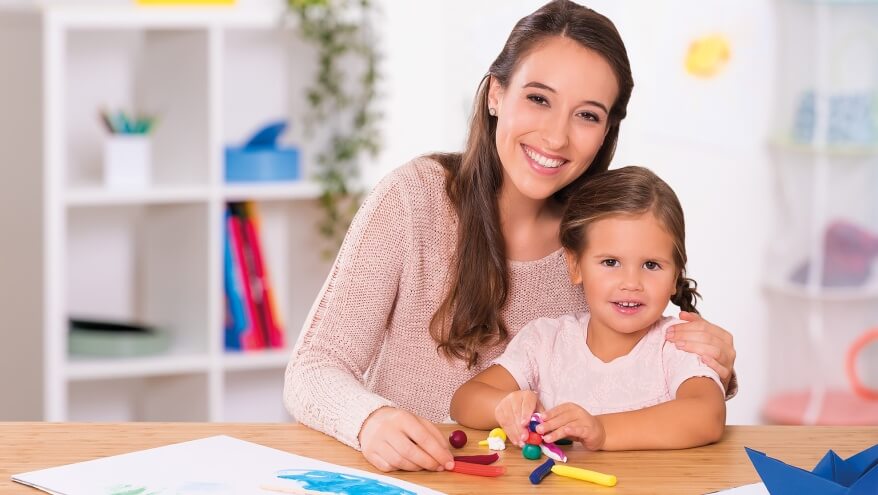 Eine Mutter und ihre Tochter lächeln in die Kamera. Sie sitzen an einem Holztisch mit Bastelmaterial und Wachsmalstiften von Pelikan. Im Hintergerund sieht man ein Regal mit verschiedenen Büroutensilien. Vor den beiden liegt ein Blatt Papier, auf das gemalt wurde und ein Papierschiffchen.