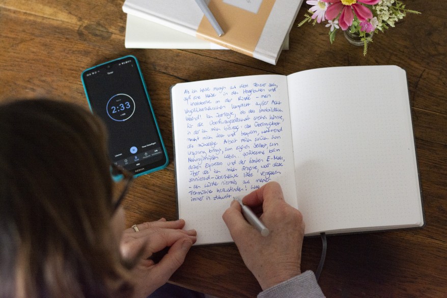 Frau sitzt am Schreibtisch und schreibt mit der Technik Free Writing in einen Text in ein Notizbuch von Hahnemühle. Ein Timer auf dem Smartphone stoppt die Zeit.