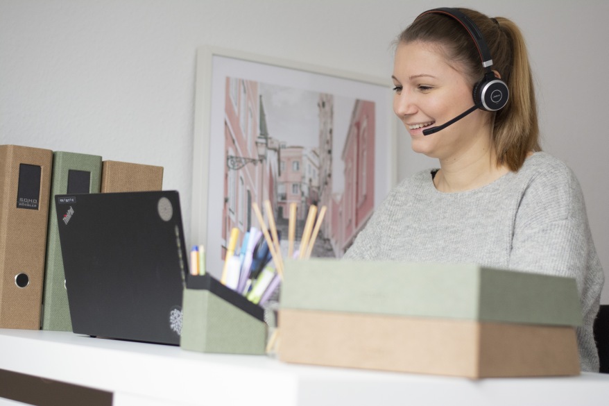 Frau sitzt am Schreibtisch und arbeitet im Homeoffice. Vor ihr steht ein aufgeklappter Laptop, sie hat ein Headset auf und ist von Büromaterialien aus der S.O.H.O.-Serie von Rössler umgeben.