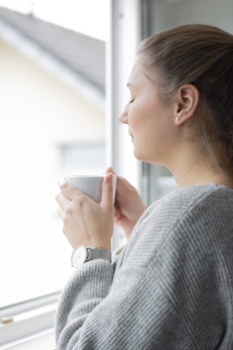 Frau steht mit geschlossenen Augen und Kaffeetasse am Fenster und macht eine Pause.