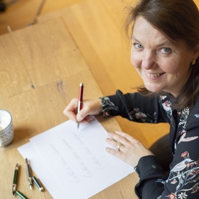 Frau sitzt am Schreibtisch und schreibt "Endlich schön schreiben!" auf ein Blatt Papier. Sie schaut zur Kamera und lächelt.