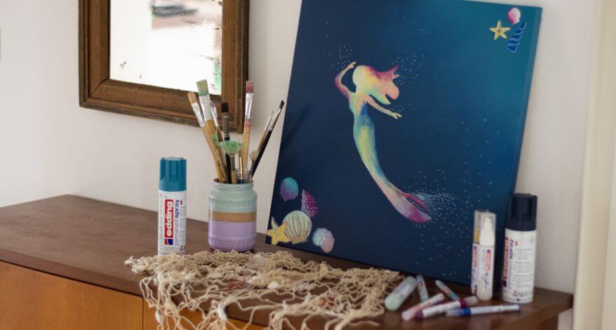 Eine mit Acrylfarben gemalte Meerjungfrau auf Leinwand steht auf einer Anrichte. Davor stehen Sprühfarben und Acrylfarben von Edding und ein Glas mit Pinseln.