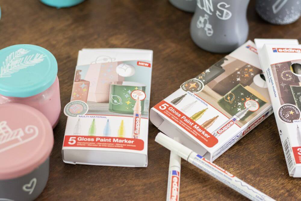 Nahaufnahme von drei Packungen Glanzlackmarkern von Edding in verschiedenen Farben, mit deren Hilfe ein Upcycling-Adventskalender aus leeren Marmeladen- und Einmachgläsern gestaltet wird.