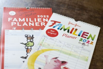 Zwei Familienplaner von Heye liegen auf einem Tisch: Familienplaner "Simons Katze" und Familienplaner "Helme Heine".in unterschiedlichsten Designs