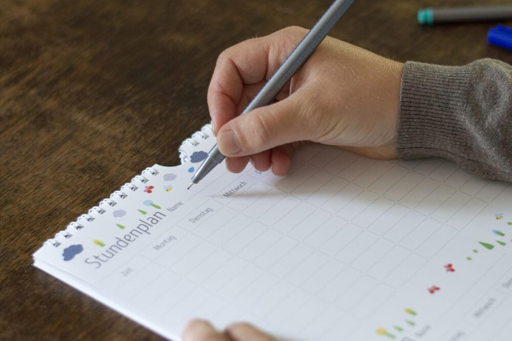 Nahaufnahme einer Hand, die in einen Familienplaner von Heye einen Stundenplan beschreibt.