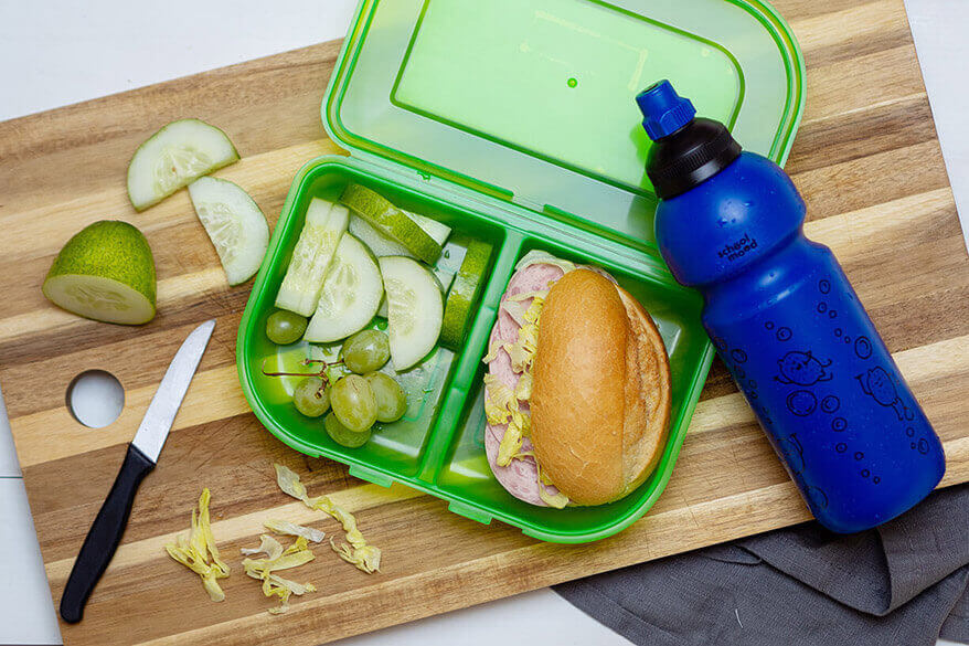 Auf einem Holzbrettchen liegt eine aufgeklappte Brotdose und eine Trinkflasche der Marke School-Mood. Die Snackbox ist mit einem belegten Brötchen, Gurkenscheiben und Trauben gefüllt. Foto: Johanna Rundel
