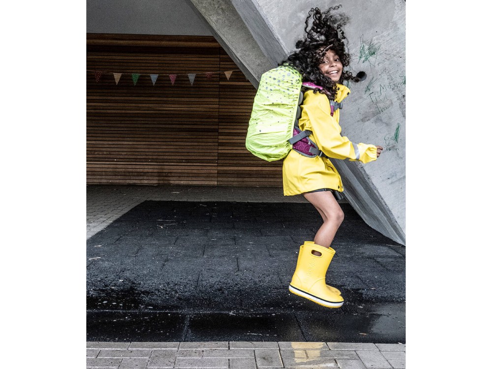 Ein Mädchen im Grundschulalter hüpft in gelben Gummistiefeln und gelber Regenjacke vor einem Schulgebäude in eine Pfütze. Sie trägt dabei einen Schulranzen der Firma School-Mood und eine dazu passende Regenhaube.