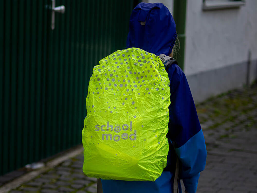 Ein Grundschulkind in blauer Regenjacke mit aufgezogener Kapuze läuft eine regnerische Straße entlang. Die neongelbe Schulranzen-Regenhaube von School-Mood leuchtet gut sichtbar in der Dunkelheit. Foto: Johanna Rundel