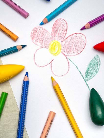 Auf einem weißen Blatt Papier ist eine Blume gemalt. Drumherum liegen verschiedene Jumbo Grip Buntstifte sowie radierbare Malkreide in Birnenform der Marke Faber-Castell. Foto: Johanna Rundel