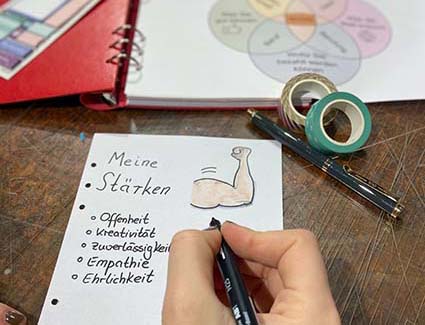 Eine Hand hält einen schwarzen Marker und schreibt auf ein Blatt Papier auf dem "Meine Stärken" steht. Im Hintergrund sieht man ein aufgeschlagenes Notizbuch und Washitape.
