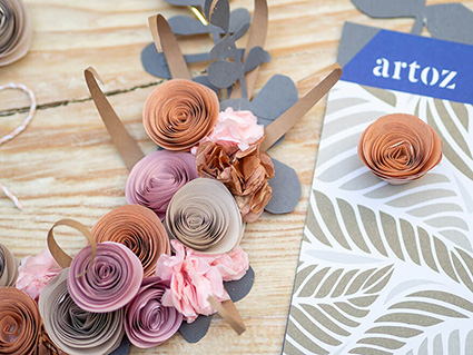 Auf einer hellen Holzoberfläche liegen ein goldener Kranz mit verschiedenen Papierblumen. Am rechten Bildrand befinden sich eine Packung Papier von Artoz. Foto: Johanna Rundel