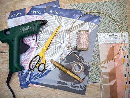 Auf einer hellen Holzoberfläche liegen alle Materialien, die man zur Herstellung eines Papierblumenkranzes benötigt. Kartons verschiedener Grammaturen und Seidenpapier von Artoz, ein goldener Metallring, eine Heißklebepistole, goldener Draht, eine goldene Schere und Bäckergarn. Foto: Johanna Rundel