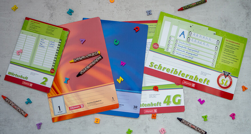 Mehrere Schulhefte liegen nebeneinander, darüber Wachsmalstifte und kleine Buchstaben aus Gummi.