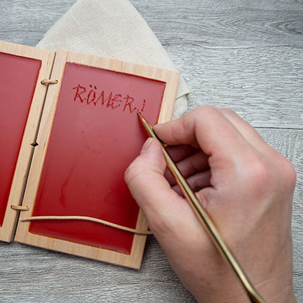 Auf einem rustikalen Holzuntergrund liegt ein kleiner Jutebeutel, auf dem eine aufgeklappte römische Wachstafel mit rotem Wachs liegen. Jemand schreibt mit einem Griffel aus Bronze das Wort "Römer!" in das Wachs. Foto: Johanna Rundel