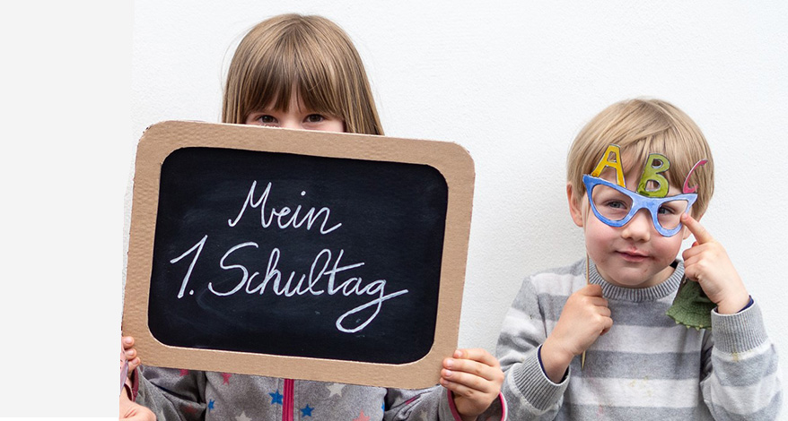 Links hält ein Mädchen im Grundschulalter, mit blonden Haaren und roten Kleidern, einen Foto-Prop Bilderrahmen aus braunem Pappkarton, mit verschiedenen Schulmaterialien aus Papier (Lineal, Stift, Buch etc.) darauf, vor sich in die Kamera. In der Mitte steht ein braunhaariges Mädchen im selben Alter und hält sich eine kleine Schultafel vor das Gesicht, auf der "Mein 1. Schultag" steht. Rechts daneben steht ein kleiner Junge mit einem Foto-Prop in Form einer Brille, mit den Buchstaben ABC darauf, vor den Augen. Der für die Props verwendete Fotokarton ist von der Marke Heyda, die Holzstäbe sind von Knorr-Prandell. Foto: Johanna Rundel