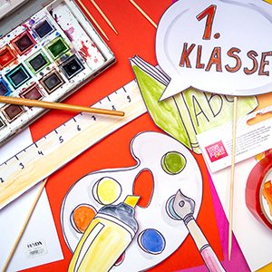 Verschiedene Fotoprops für Einschulungsfotos liegen auf einem roten Untergrund. Zu sehen sind unter anderem: eine Sprechblase, in der "1. Klasse" steht, eine blaue Brille, ein bunter Wasserfarbkasten, ein Buntstift und ein gelber Textmarker. Der für die Props verwendete Fotokarton ist von der Marke Heyda, die Holzstäbe sind von Knorr-Prandell. Foto: Johanna Rundel