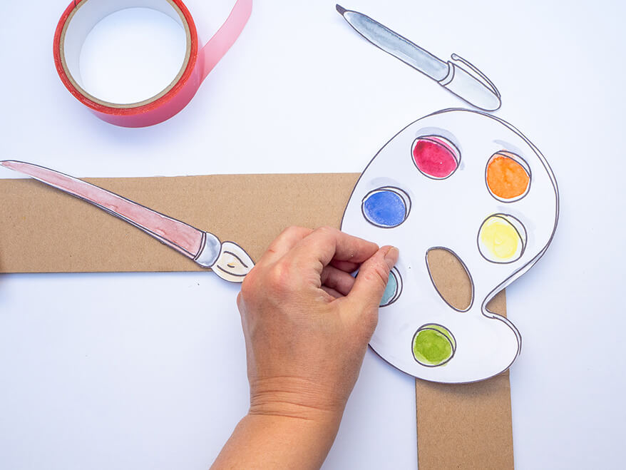 Jemand möchte Foto-Props für die Einschulung basteln und klebt dafür eine ausgeschnittene und bemalte Farbpalette auf einen großen Bilderrahmen aus brauner Pappe. Foto: Johanna Rundel
