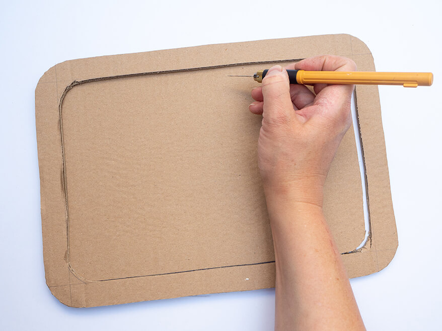 Jemand schneidet mit einem Cutter aus einer braunen Pappe einen Rahmen um daraus eine kleine Schultafel als Foto-Prop für die Einschulung zu basteln. Foto: Johanna Rundel