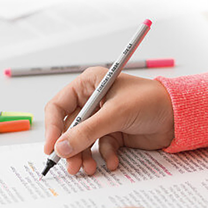 Jemand hält einen Stift von Pelikan im Dreipunktgriff über einem Blatt mit Wörtern. Im Hintergrund sind weitere verschiedenfarbige Stifte zu sehen. Die Person hat einen pinken Pulli an.