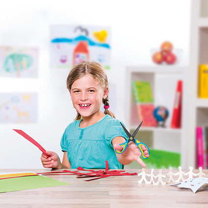 Ein Mädchen hält in der linken Hand eine blaue Schere von Pelikan. In der rechten Hand hält sie einen roten Bogen Papier. Sie sitzt an einem Tisch an dem verschiedene bunte Bastelpapiere liegen. In der rechten Ecke steht ein Wimpel aus ausgeschnittenen Männchen. 