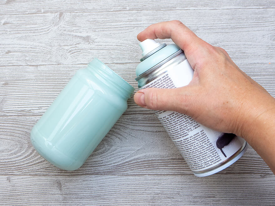 Jemand sprüht auf einem hellgrauen Holzuntergrund ein leeres Marmeladenglas von innen mit hellblauer Acrylfarbe aus.