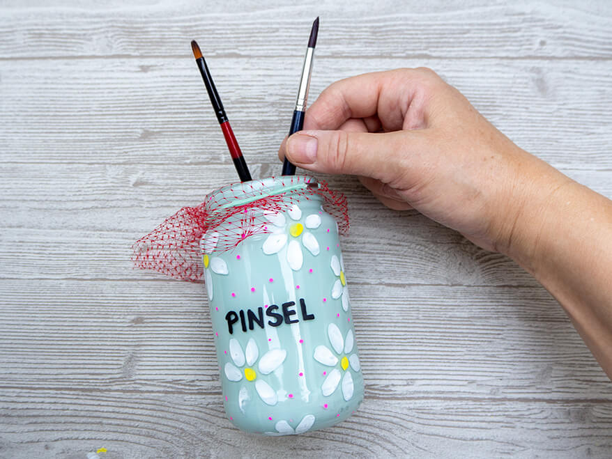 Jemand steckt auf einem hellgrauen Holzuntergrund Pinsel in ein leeres Marmeladenglas, das von innen mit hellblauer Acrylfarbe ausgesprüht wurde und mit Acrylmarkern angemalt wurde, mit einem roten Netz über der Öffnung.