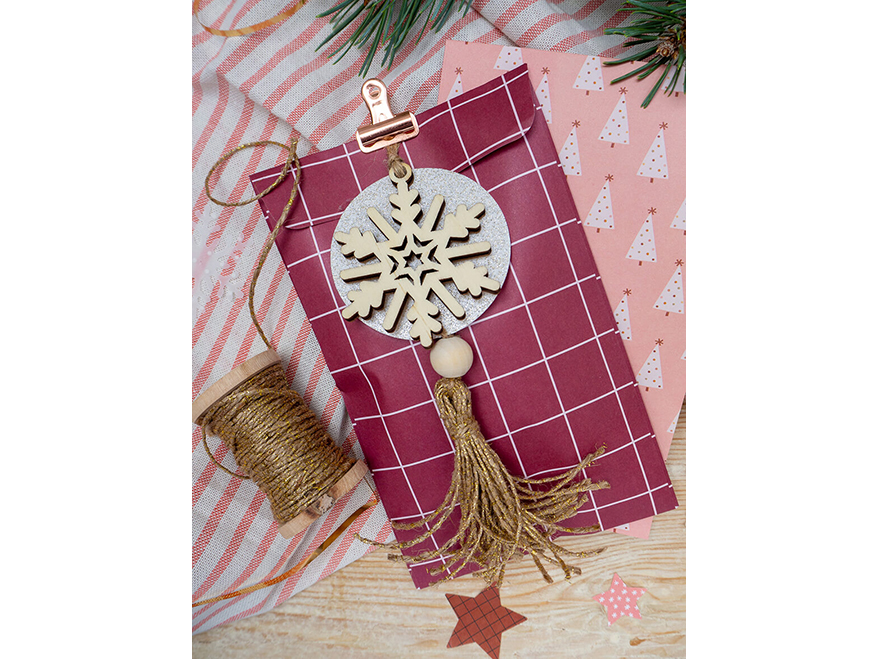 Eine rot-weiß karierte Geschenktüte liegt auf einem Holzbrett, auf dem ein weiß-rosa gestreifte Decke drapiert ist. An der Geschenktüte wurde ein Geschenkanhänger befestigt. Er besteht aus einem Papierkreis, auf dem eine Holzschneeflocke liegt und einer goldenen Tassel. Außerdem ist eine Holzrolle mit goldener Kordel, Sternenaufkleber und ein Tannenzweig zu sehen. Foto: Johanna Rundel