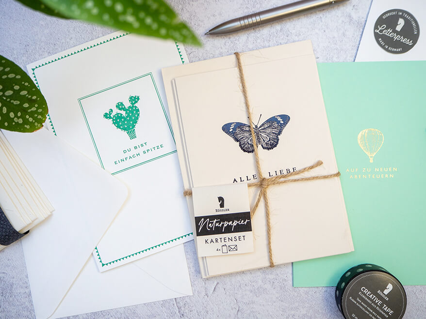 Auf einem hellgrauen Untergrund liegen verschiedene Letterpress-Grußkarten in verschiedenen Grüntönen von Rössler. Daneben sieht man ein mit Jutefaden zusammengebundenes Bündel Grußkarten aus Naturpapier. Drumherum sind ein silberner Kugelschreiber, eine Rolle schwarzes Washitape mit weißen Punkten, sowie ein weiteres Bündel mit Briefumschlägen zu sehen. Aus der linken oberen Ecke des Bildes ragen die Blätter einer Zimmerpflanze ins Bild. Foto: Johanna Rundel