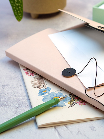 Auf einem hellgrauen Untergrund liegen ein Notizheft mit Blumen sowie eine rosa Sammelmappe von Rössler. Die Mappe ist leicht geöffnet und man sieht verschiedene Schreibpapiere herausschauen. Auf dem Heft liegt ein hellgrüner Füller. Im Hintergrund ist eine Zimmerpflanze zu sehen. Foto: Johanna Rundel