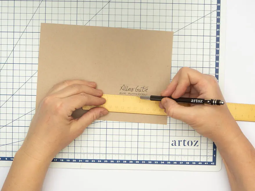 Jemand schneidet mit einem Cutter Schlitze ober- und unterhalb eines Letterings in eine Klappkarte aus Kraftkarton von Artoz. Der Untergrund ist weiß. Foto: Johanna Rundel