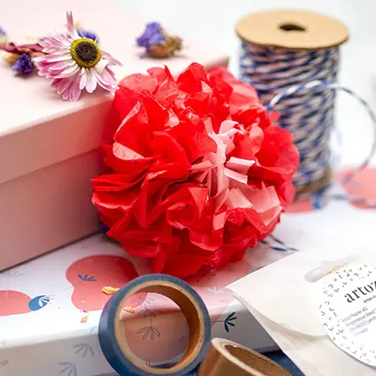 Verschiedene Materialien der Marke Artoz zum Verpacken von Geschenken: Geschenkschachtel aus Pappe, Washitape, Blütenkonfetti, Geschenkanhänger, eine rote Blume aus Seidenpapier, Trockenblumen und Bäckergarn, eine goldene Schere, Konfetti, Geschenkanhänger und Trockenblumen. Foto: Johanna Rundel
