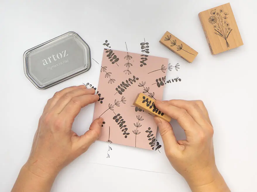 Jemand stempelt botanische Motive (Eukalyptuszweige und Gräser) auf eine altrosa Klappkarte von Artoz. Das Stempelkissen sowie weitere Stempel liegen ebenfalls im Bild. Der Untergrund ist weiß. Foto: Johanna Rundel