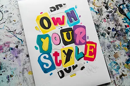 Auf einem mit Farbe beklecksten Untergrund liegt ein weißes DIN A4 Blatt mit einem Lettering in bunten Druckbuchstaben.