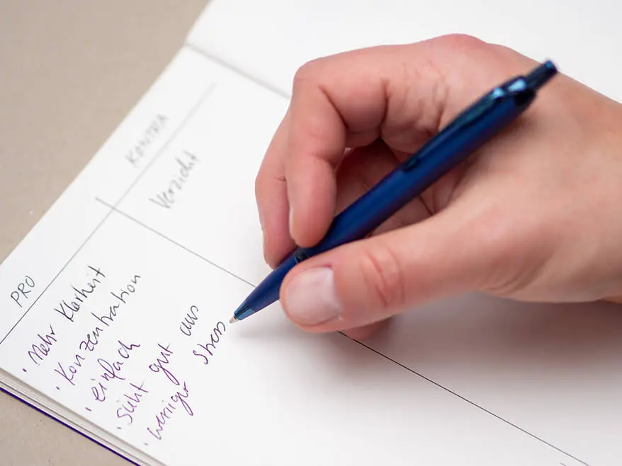 Jemand schreibt mit einem blauen Kugelschreiber von Parker eine Pro- und Kontraliste in ein Notizbuch. Foto: Johanna Rundel