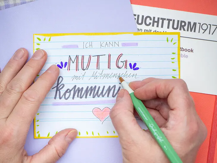 Jemand schreibt mit einem Drehgriffel von LEUCHTTURM1917 den Spruch „ich kann mutig mit meinen Mitmenschen kommunizieren“ auf eine Affirmationskarte. Foto: Johanna Rundel