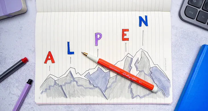 Auf einem grauen Schreibtisch liegt ein aufgeschlagenes Notizheft von LEUCHTTURM1917 in das eine Berglandschaft gemalt wurde. Darüber stehen über jeder Bergspitze je ein Buchstabe des Worts „ALPEN“. Foto Johanna Rundel