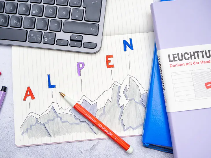 Auf einem grauen Schreibtisch liegt ein aufgeschlagenes Notizheft von LEUCHTTURM1917 in das eine Berglandschaft gemalt wurde. Darüber stehen über jeder Bergspitze je ein Buchstabe des Worts „ALPEN“. Foto Johanna Rundel