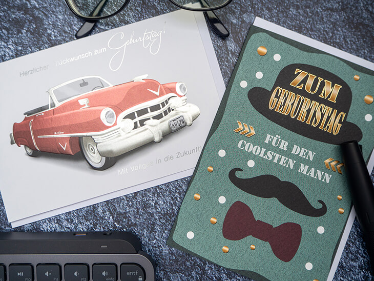 Zwei Geburtstagskarten von ABC Grußkarten mit männlichen Motiven liegen auf einem dunklen Schreibtisch. Foto: Johanna Rundel