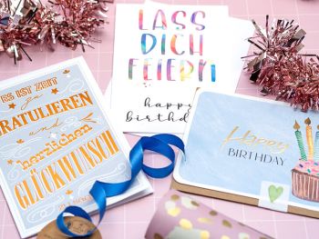 Drei verschiedene, bunte Geburtstagskarten von ABC Grußkarten liegen auf einem rosa Untergrund zwischen diversen Geburtstags-Accessoires. Foto: Johanna Rundel