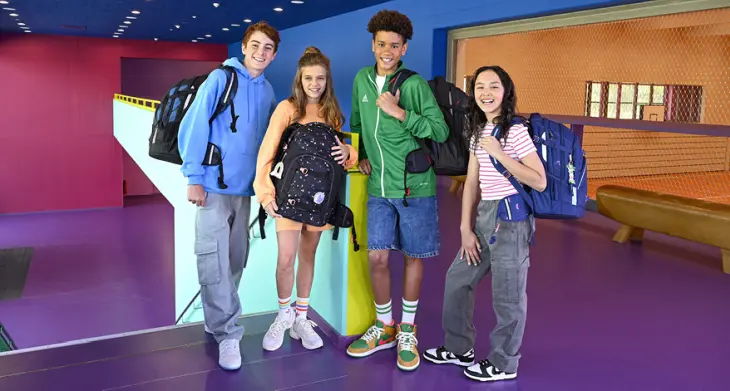 Vier Jugendliche stehen in einem bunten Raum, tragen Schulrucksäcke von coocazoo und lachen in die Kamera.