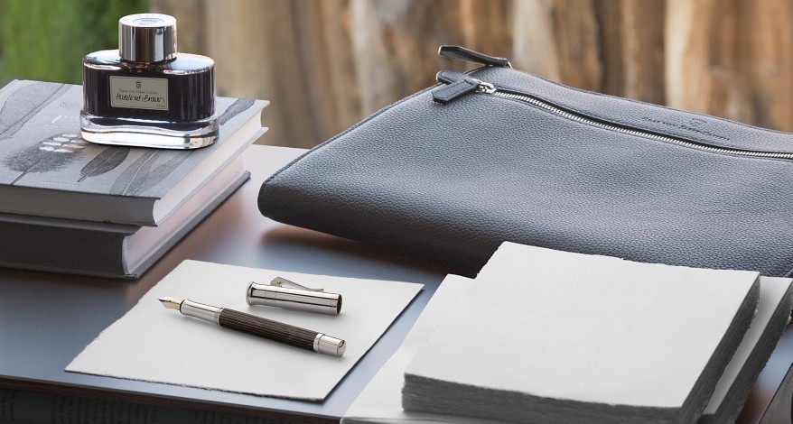 Auf einem Tisch liegen ein Stift, Tinte und eine Ledermappe von Graf von Faber-Castell, Papierstapel und 2 Bücher auf dem Tisch