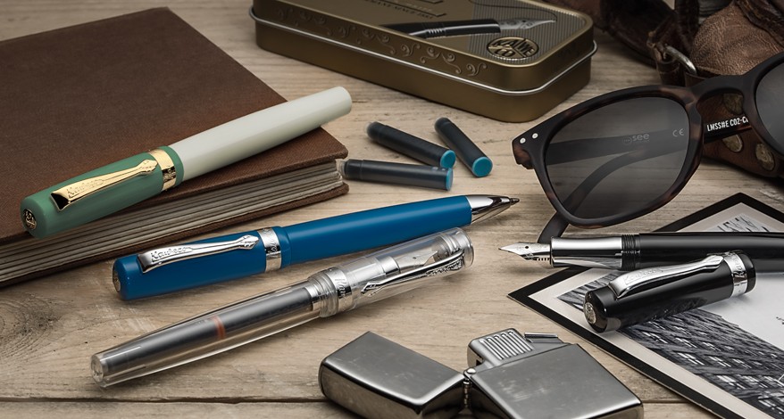 Verschiedene Kaweco-Stifte auf Schreibtisch mit Sonnenbrille und Notizbuch.