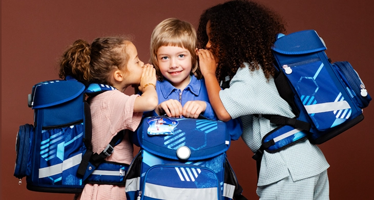 Zwei Kinder mit Schulranzen von School-Mood auf dem Rücken, flüstern einem Kind ins Ohr, welches auch einen Schulranzen in der Hand hält