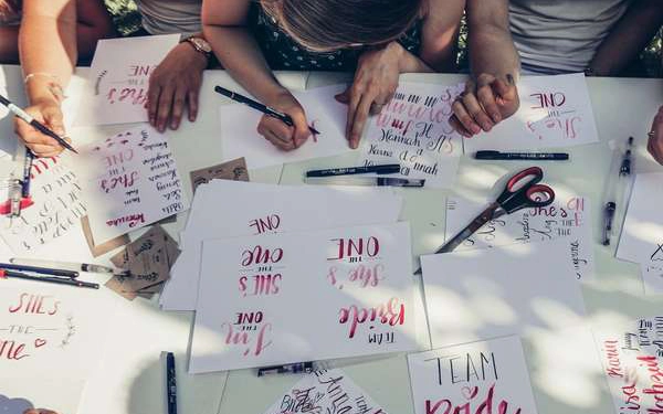 Ein Tisch mit zahlreichen beletterten Blättern. Am oberen Bildrand lettern einige Personen Sprüche und Worte auf Blätter mit Hilfe von Brushpens.