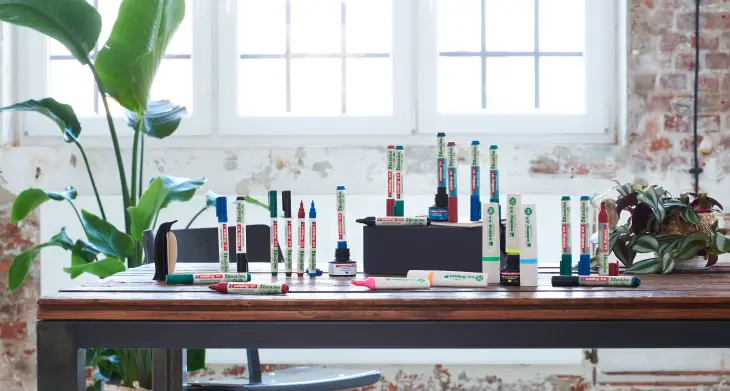 Auf einem Holzschreibtisch, in einem Raum mit großen Fenstern und Backsteinoptik, stehen verschiedene bunte edding Marker der Serie EcoLine, Auf dem Schreibtisch steht eine schwarze Kiste mit weiteren Markern darauf und eine Topfpflanze. Eine weitere Grünpflanze befindet sich im Hintergrund.