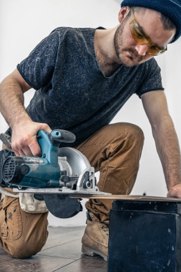Handwerker mit blauer Mütze und Schutzbrille schneidet mit einer Makita Handkreissäge ein Holzbrett. Er trägt ein dunkles T-Shirt und Arbeitshose und kniet auf dem Boden.