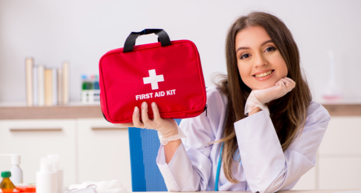 Das Bild zeigt eine lächelnde Frau in einem weißen Kittel, die eine rote Erste-Hilfe-Tasche mit einem weißen Kreuz darauf hochhält. Sie sitzt an einem Tisch, auf dem verschiedene medizinische Utensilien und Fläschchen zu sehen sind. Die Szene betont die Wichtigkeit, auf Notfälle vorbereitet zu sein.