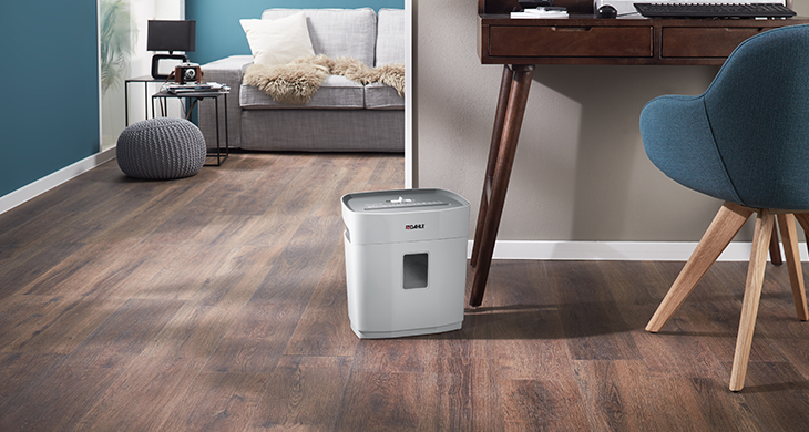 Das Bild zeigt einen kompakten Dahle Aktenvernichter in einem modernen Homeoffice-Setup mit einem Holzfußboden, einem Schreibtisch und einem blauen Stuhl im Vordergrund. Im Hintergrund ist eine gemütliche Sitzgelegenheit zu sehen.“