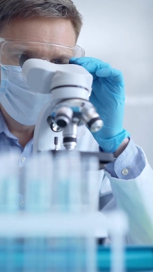 Wissenschaftler mit Schutzbrille, Atemschutzmasken und Einweghandschuhen untersucht Proben unter einem Mikroskop.