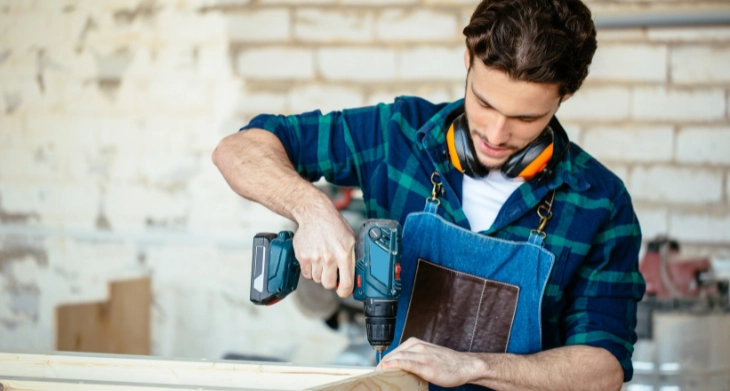 Handwerker montiert ein Holzstück mit einem Makita Akkuschrauber in einer Werkstatt. Er trägt eine blaue Schürze und Gehörschutz um den Hals.
